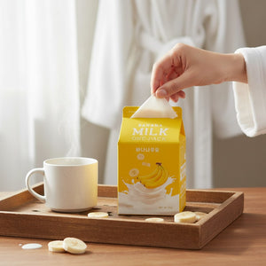 Skin TOGO A'pieu Milk One Pack banana sheet mask being pulled from its yellow carton on a wooden breakfast tray.