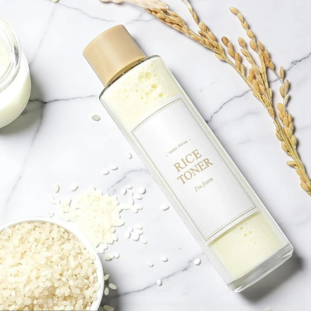 Skin TOGO I'm from Rice Toner in a clear bottle with milky liquid, surrounded by raw rice grains on a marble surface.